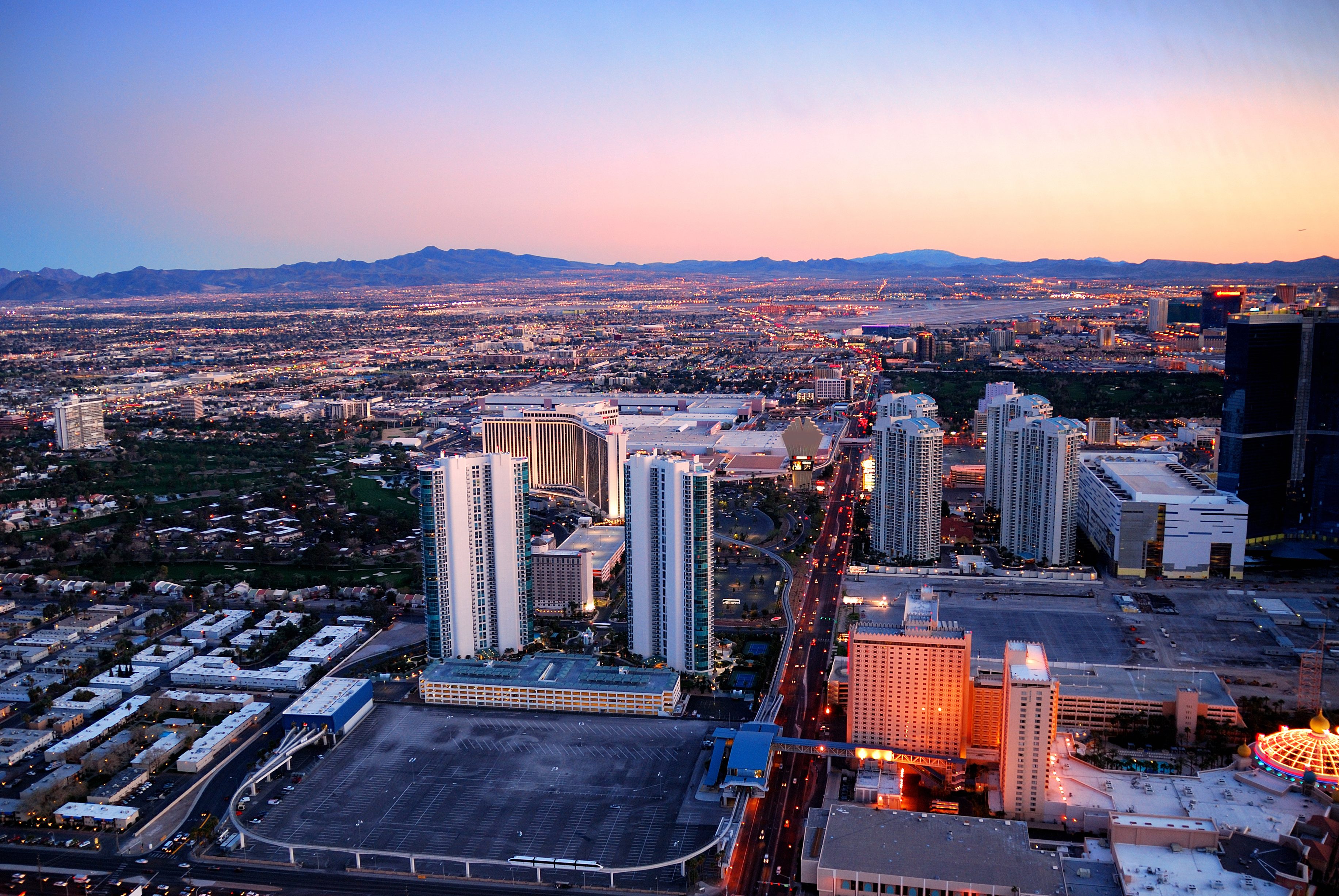 Aerial view of a vibrant cityscape at sunset, showcasing tall buildings, city lights, and expansive mountains in the background.